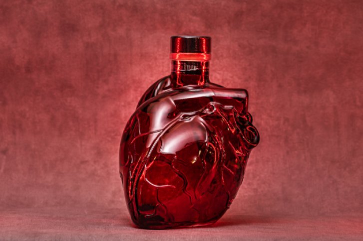 Rote herzförmige Flasche steht auf rotem Hintergrund, reflektiert Licht, ohne sichtbaren Inhalt.