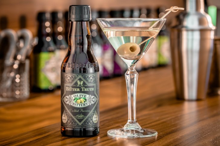 Bitter-Flasche steht neben Martini-Glas mit Olive, auf Holztresen, Barhintergrund mit Flaschen.