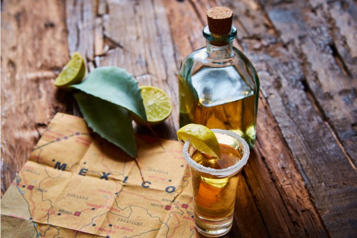 Tequila-Glas mit Limette steht neben Flasche auf Holztisch, Karte von "MEXICO" darunter.