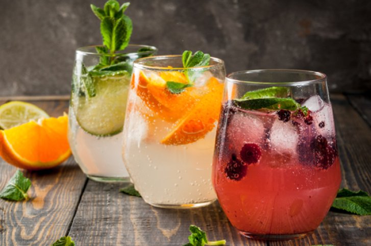 Drei farbige Cocktails mit Früchten und Minze stehen auf einem Holztisch vor grauem Hintergrund.