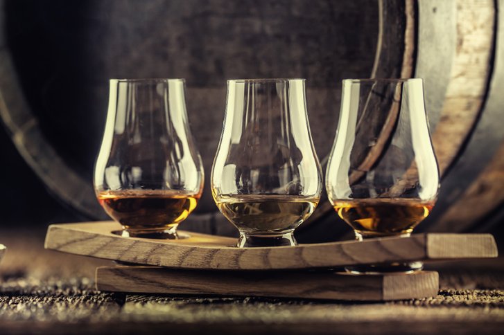 Drei Whiskygläser stehen auf einem Holzbrett, im Hintergrund ein großes Holzfass.