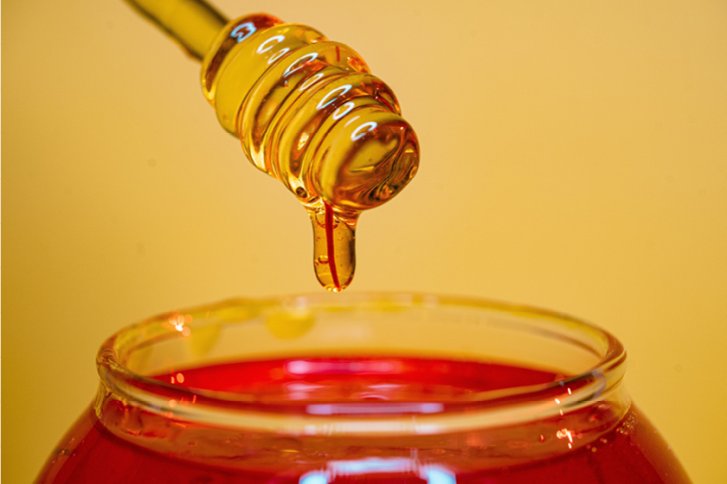 Honiglöffel tropft goldenen Honig in ein Glas auf gelbem Hintergrund.