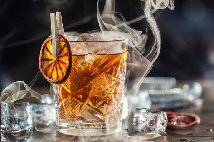 Kristallglas mit Whisky, raucht, auf Holztisch mit Eiswürfeln und getrockneter Orangenscheibe befestigt.