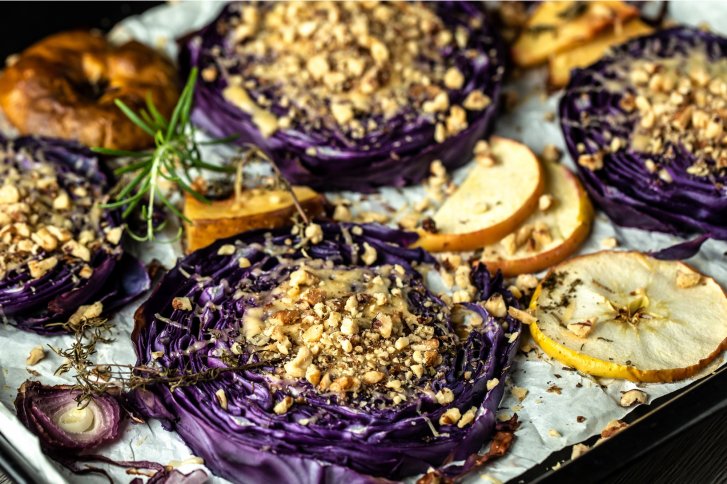 Rotkohlscheiben, mit Nüssen bestreut, liegen auf Backblech, umgeben von Apfelscheiben und Kräutern.