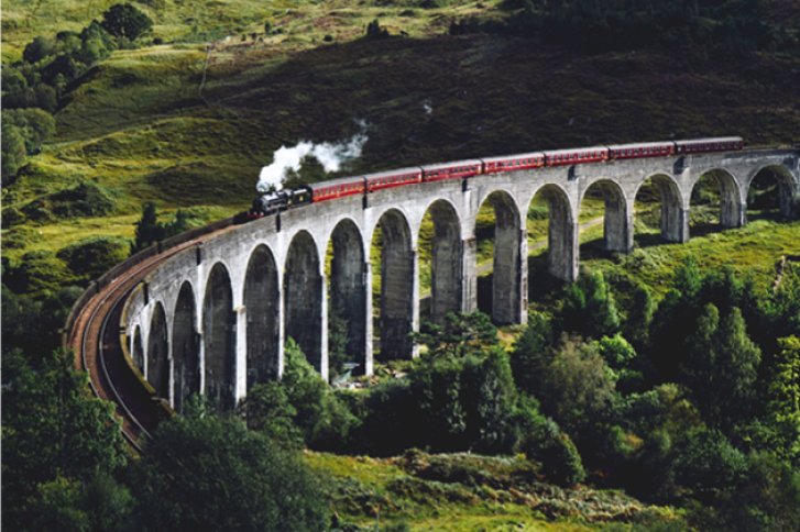 Zug fährt über steinernes Viadukt in grüner, hügeliger Landschaft. Rauch steigt aus der Lokomotive.