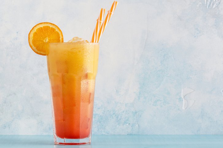 Ein hoher Cocktail mit Orangenscheibe und Strohhalmen, steht vor hellblauer Wand.