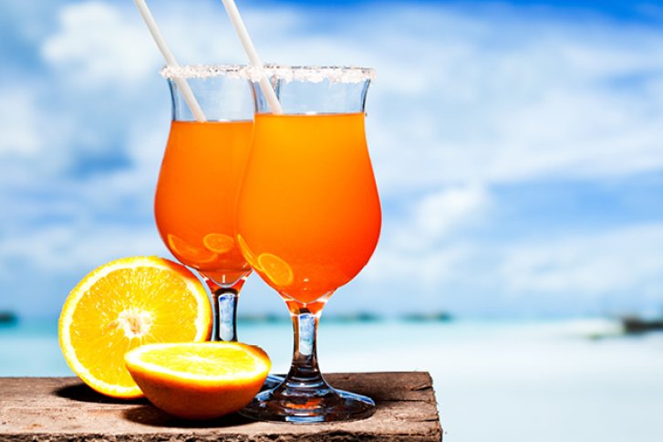 Zwei Cocktails stehen auf Holztisch am Strand. Dekoriert mit Orangenscheiben, blauer Himmel im Hintergrund.
