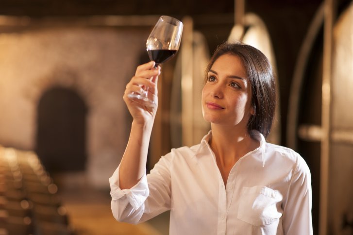 Frau betrachtet ein Weinglas sorgfältig im Lager einer Weinkellerei mit Holzfässern.