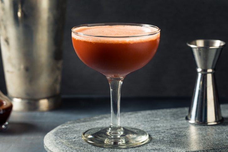 Ein Cocktailglas mit roter Flüssigkeit ruht auf einer grauen Oberfläche, umgeben von Barutensilien.