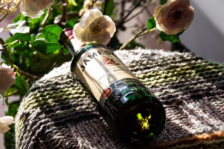 Eine Whiskyflasche liegt auf einem Strickstück, umgeben von blühenden Rosen im Sonnenlicht.