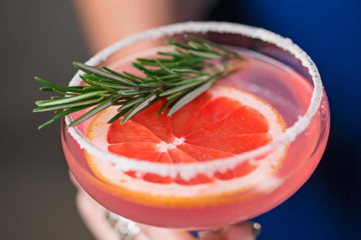 Cocktailglas enthält rosa Getränk mit Grapefruitrad, Rosmarinzweig, im Hintergrund eine verschwommene Hand.