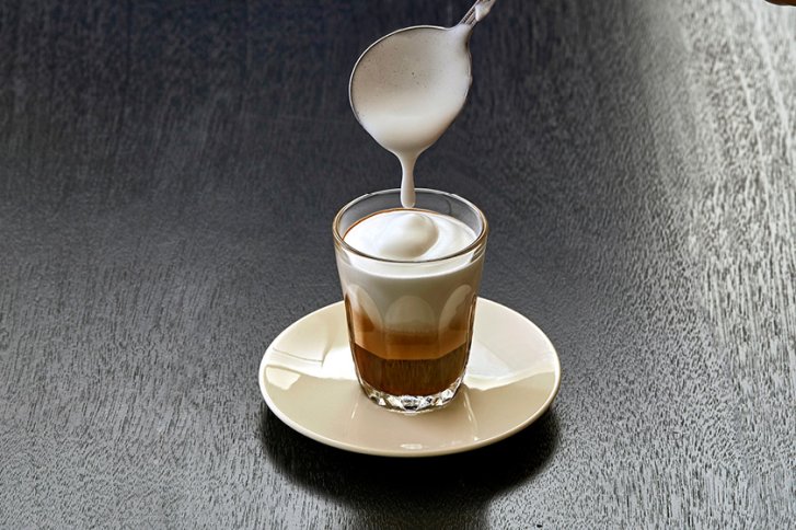 Ein Löffel gießt Schaum auf Espresso in Glas auf Untertasse auf dunklem Tisch.