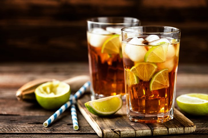 Zwei Gläser mit Eis, Limettenscheiben und dunklem Getränk auf einem Holztisch, Strohalm daneben.