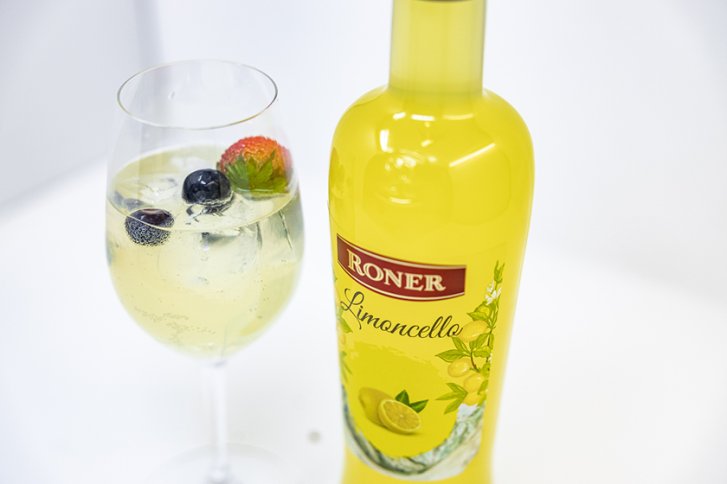 Eine gelbe Limoncello-Flasche neben einem Glas mit Eisbwürfeln und Beeren auf weißem Hintergrund.