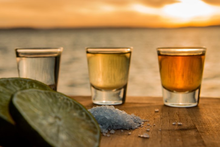 Drei Tequila-Gläser stehen auf Holztisch, umgeben von Limetten und Salz, bei Sonnenuntergang am Meer.