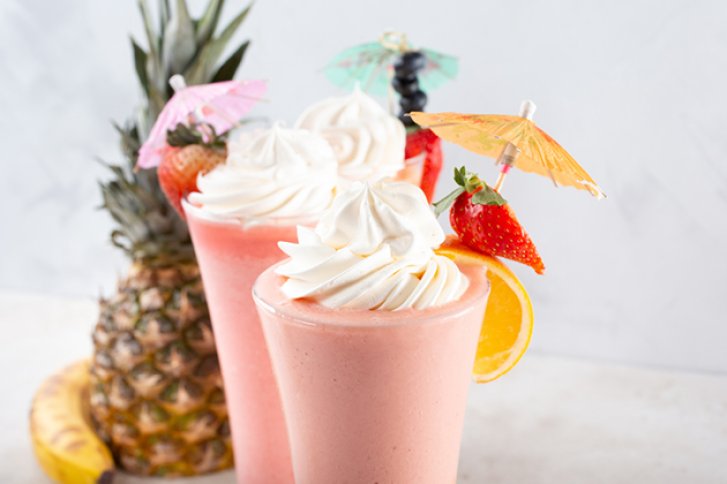 Drei Dessertgläser mit Sahne, dekoriert mit Früchten und Schirmchen, stehen neben Ananas und Banane.