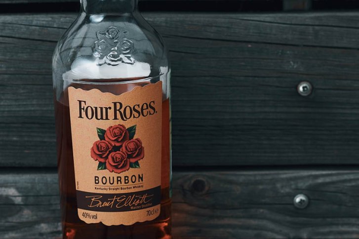 Eine Bourbon-Flasche steht vor einer hölzernen Wand, mit Etikett "Four Roses" und Rosenmotiv.