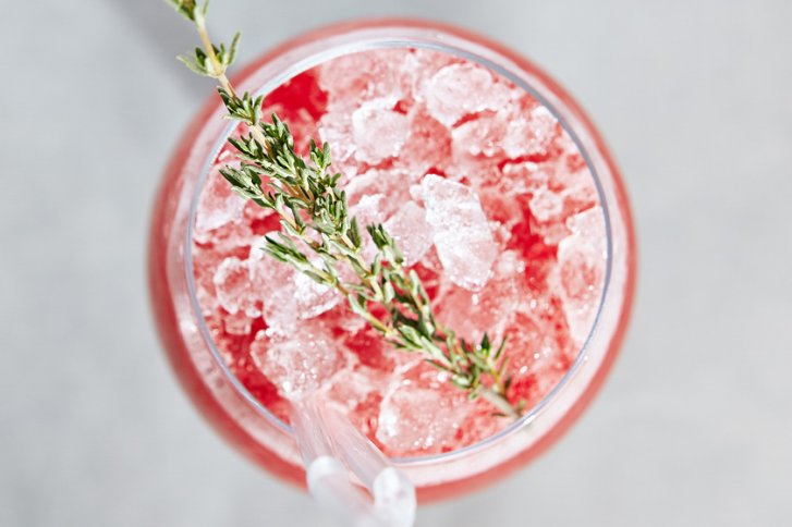 Ein rosafarbener Cocktail mit Crushed Ice und Thymianzweig, im Glas mit zwei Strohhalmen.
