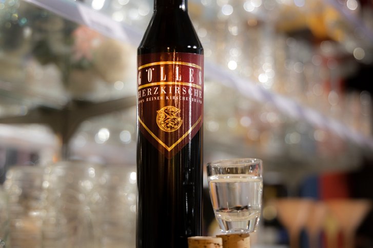 Schwarze Flasche steht mit gefülltem Schnapsglas und Korken vor einer verschwommenen Bar.