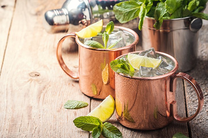 Kupferbecher stehen gefüllt mit Eis, Zitronenscheiben und Minze auf einem Holztisch. Hintergrund: Cocktailbedarf.