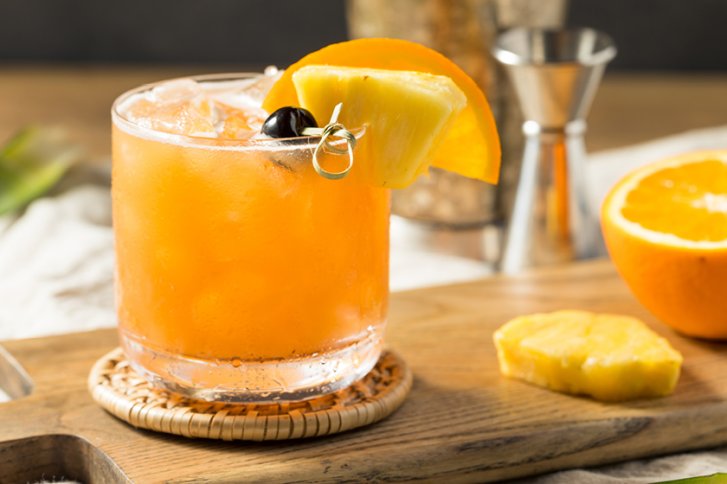 Ein Cocktailglas mit Orangensaft und Eiswürfeln steht auf einem hölzernen Untersetzer, dekoriert mit Ananas.