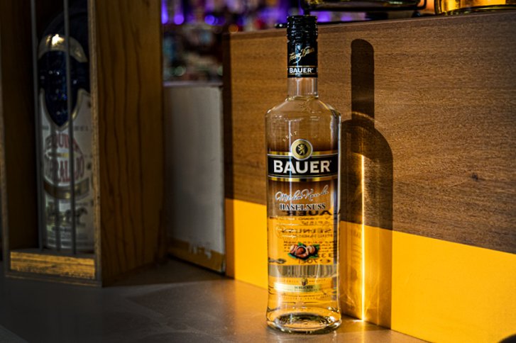 Flasche mit Aufschrift "BAUER" steht auf Holztresen, Hintergrund unscharf, andere Flasche sichtbar.