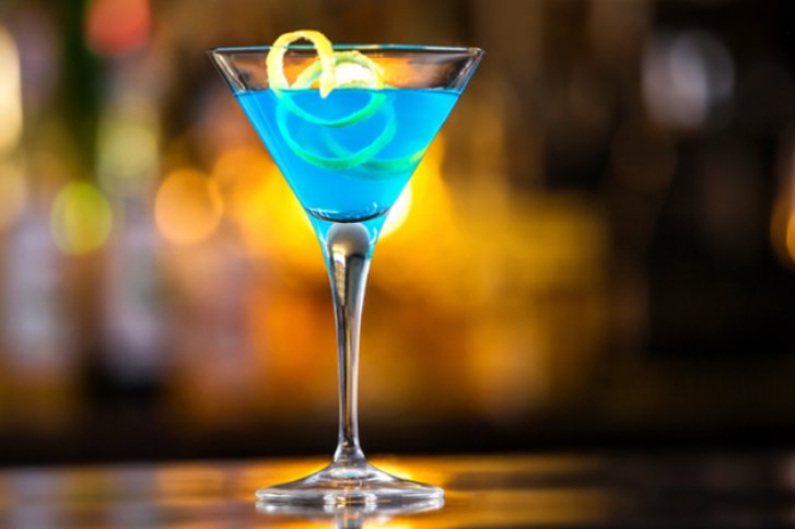 Ein blauer Cocktail in einem Martini-Glas mit Zitronenschale auf einem Thekenhintergrund.