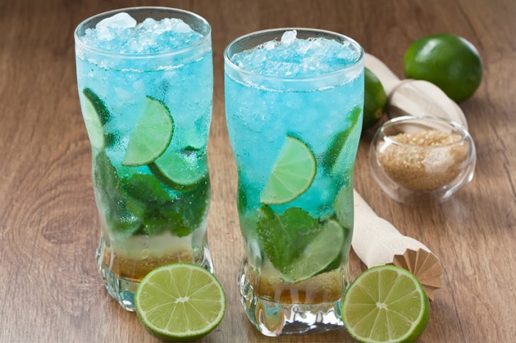 Zwei blaue Cocktails mit Limettenscheiben stehen auf einem Holztisch, umgeben von Limetten und Zucker.