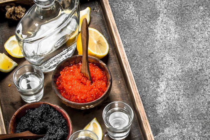 Glaskaraffe, gefüllt mit klarer Flüssigkeit, steht auf Tablett neben Kaviar, Zitronen und Schnapsgläsern.