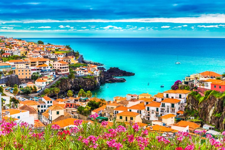 Bunte Häuser überblicken das türkisfarbene Meer, umgeben von blühenden Blumen und felsiger Küste.