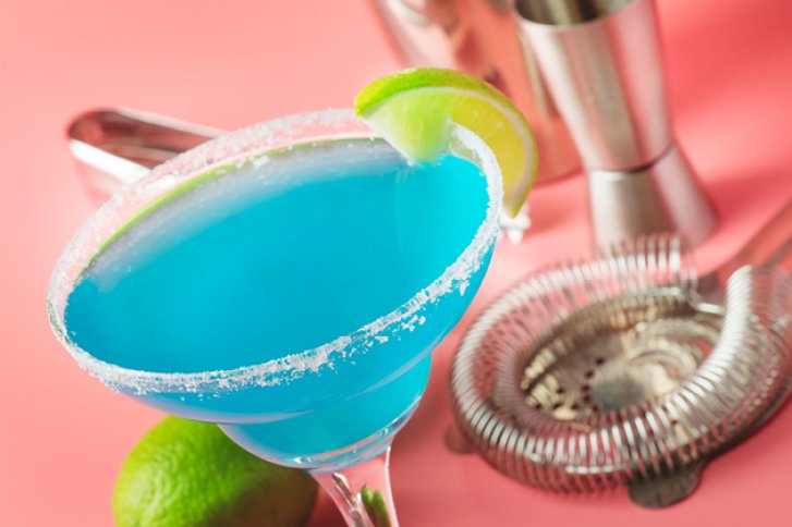 Ein blaues Cocktailglas steht mit Limettenscheibe und Salzrand vor Cocktail-Zubehör auf rosa Hintergrund.