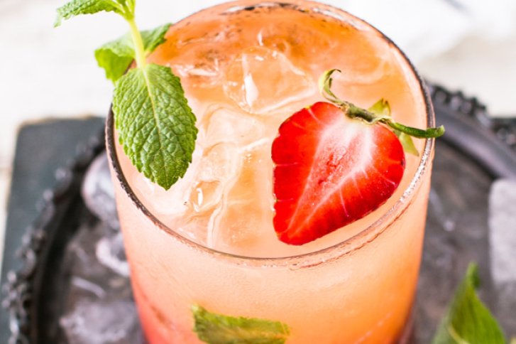 Ein Cocktailglas enthält rosafarbenes Getränk mit Eis, garniert mit Erdbeerscheibe und Minze, auf Schieferplatte.