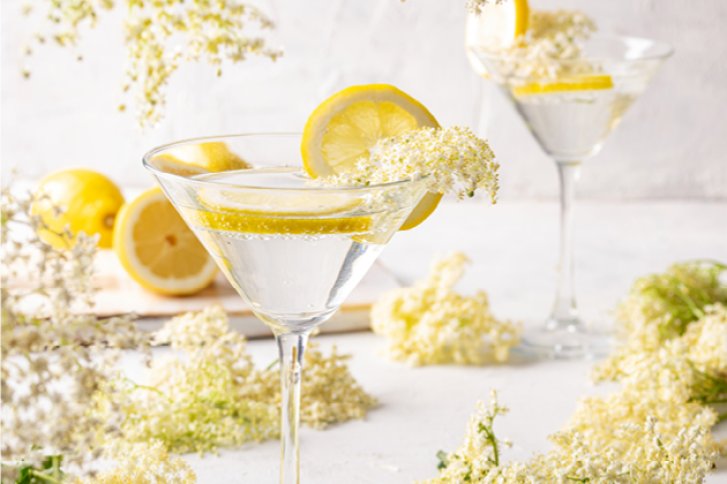 Cocktailgläser mit Zitronenscheiben und Holunderblüten dekoriert, auf weißem Tisch mit Blüten.