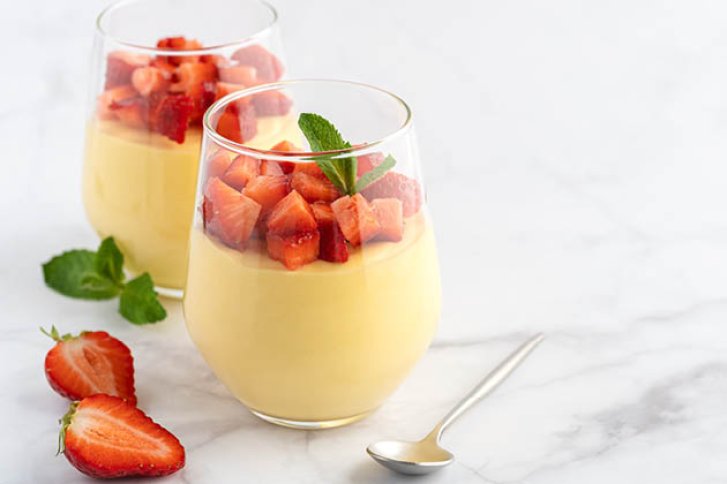 Zwei Gläser mit gelbem Dessert, garniert mit Erdbeeren, stehen auf weißem Tisch neben Löffel.