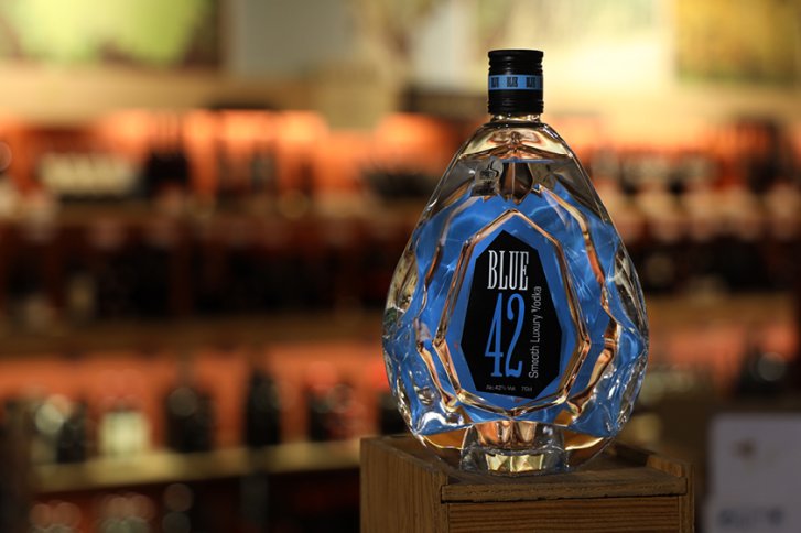 Flasche Blue 42 Vodka steht auf Holzpodest, umgeben von verschwommenen Regalen in einem Laden.
