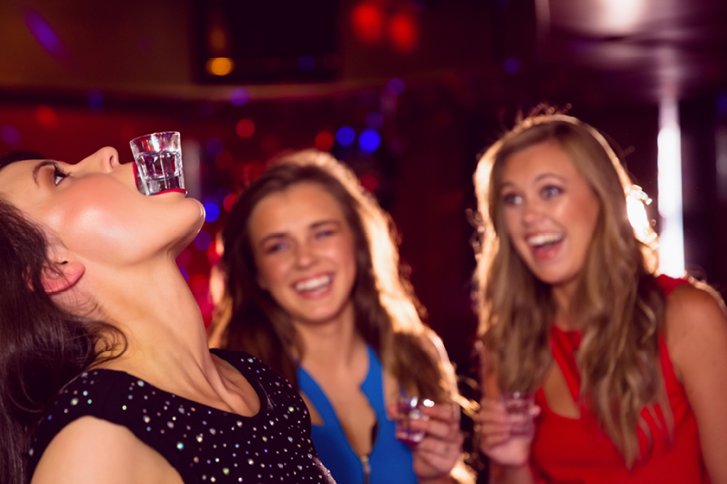 Frau balanciert Shotglas auf Kinn, während andere lachend zusehen, in einer Partyumgebung.