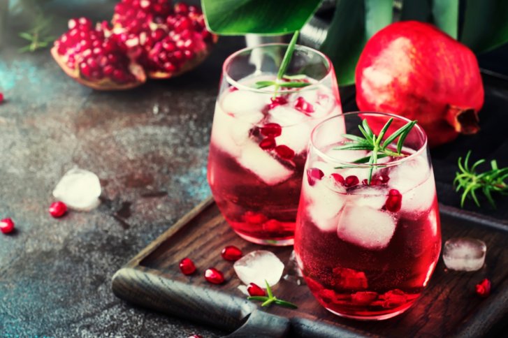 Zwei Gläser mit rosafarbenem Getränk, Granatapfelkernen und Eiswürfeln auf einem Holztisch.