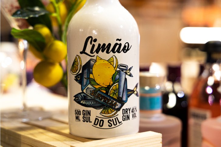 Flasche mit Zitronenmotiv steht auf Holzunterlage, Hintergrund unscharf mit weiteren Flaschen. Text: "Limão".