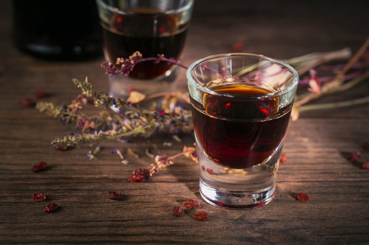 Ein Likörglas steht auf Holztisch, umgeben von getrockneten Kräutern und Beeren.