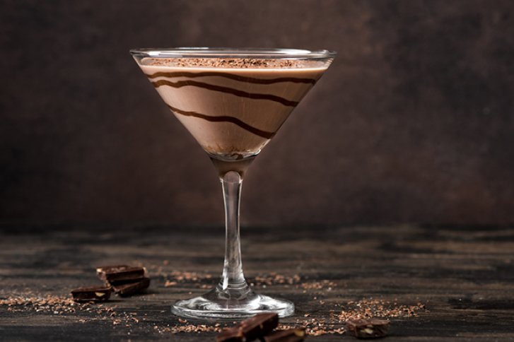 Ein Schokoladen-Martini steht dekorativ auf einem Holztisch mit Schokoladenstücken und Kakaopulver.