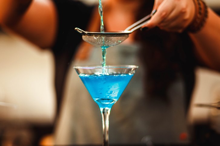 Blauer Cocktail fließt durch Sieb in Glas, gehalten von Händen, in Barumgebung.