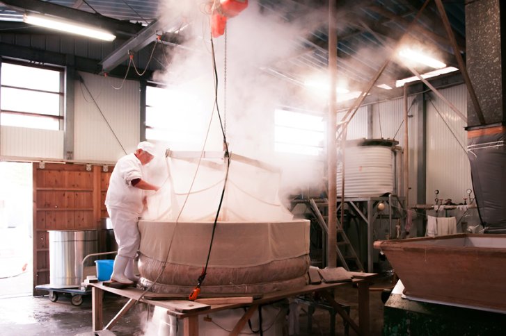 Ein Arbeiter überwacht dampfenden Bottich in heller, industrieller Produktionshalle mit großen Tanks.