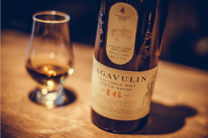 Eine Whiskyflasche und ein Glas stehen auf einem Holztisch in schummrigem Licht.