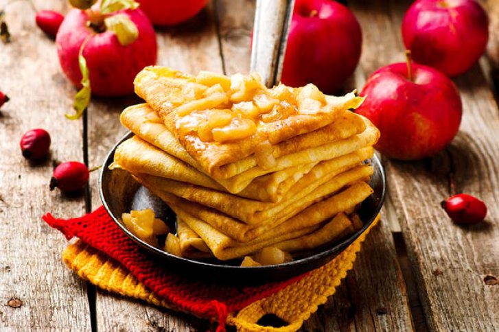 Pfannkuchen mit Apfelstücken liegen in einer Pfanne, umgeben von roten Äpfeln auf Holztisch.