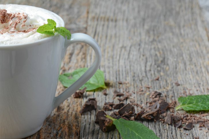 Tasse mit Sahne und Minze, steht auf Holztisch mit Schokoladenstückchen und Minzblättern.