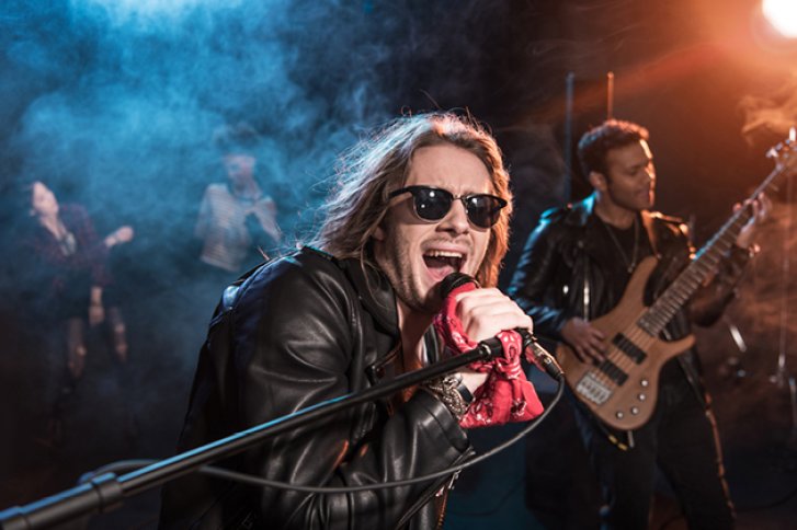 Sänger trägt Lederjacke, singt leidenschaftlich ins Mikrofon, während Band im Hintergrund spielt.