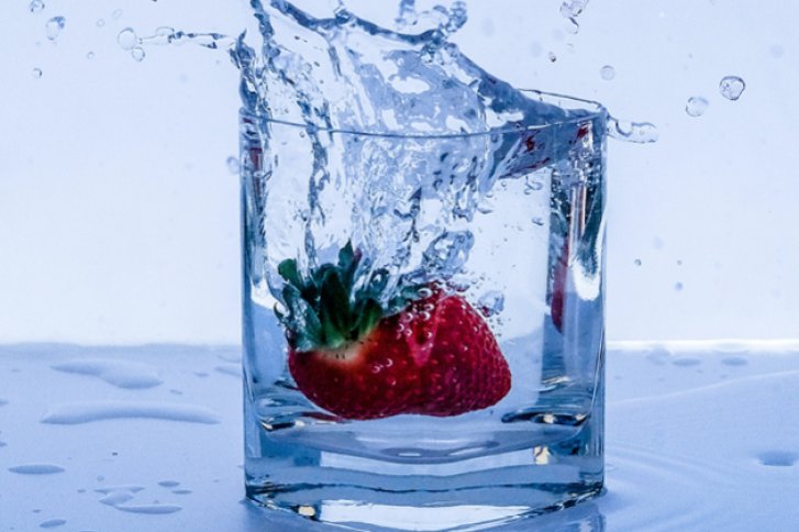 Erdbeere taucht ins Wasser, verursacht Spritzer im Glas, auf einer nassen Oberfläche.