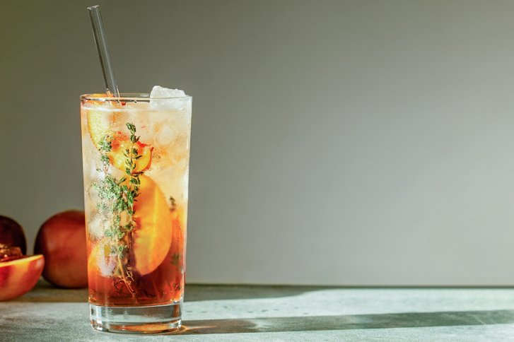 Ein Glas mit Eistee, Eiswürfeln und Pfirsich, steht auf einem Tisch in heller Umgebung.