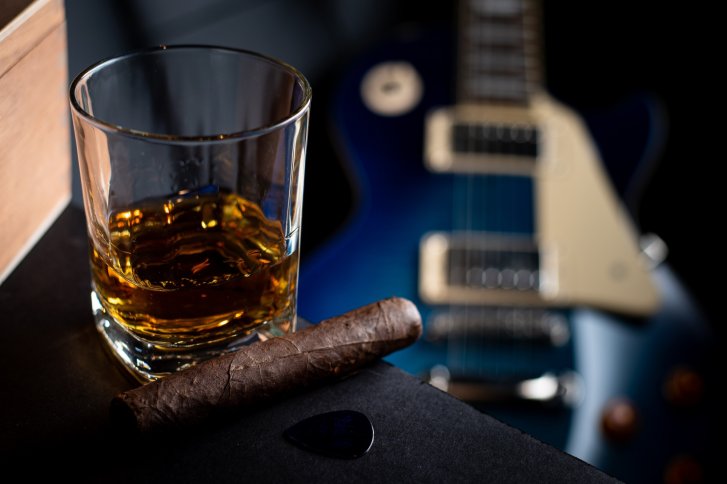 Ein Whiskeyglas steht neben einer Zigarre vor einer verschwommenen blauen E-Gitarre.