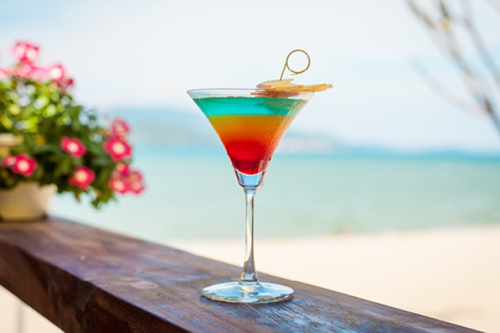Bunter Cocktail steht auf Holzgeländer, vor sandigem Strand und blauem Meer im Hintergrund.